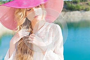 Beautiful sweet gentle girl in white dress in light big pink hat on the shore of the blue sea at sunset summer evening sun