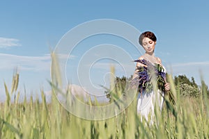 Beautiful sweet girl with a plait hair in a white summer sundress walking in a field with a bouquet of purple lupine flowers