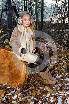 Beautiful sweet little girl dressed in a sheepskin coat and boots sitting on the trunk and in the background
