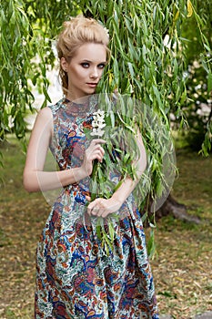 Beautiful sweet tender girl blonde with blue eyes stands near a tree with long branches with a sprig of flowers in her hands