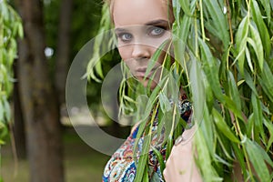 Beautiful sweet tender girl blonde with blue eyes stands near a tree with long branches with a sprig of flowers in her hands