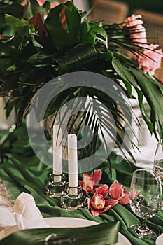 A beautiful table for a dinner party, decorated with leaves of a palm tree, white candles and flowers of a orchid