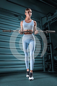 Beautiful tall blonde posing in the gym with a barbell in her hands against the background of the wall bar. The concept of sports