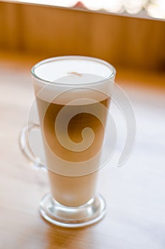 Beautiful tall transparent glass containing latte coffee drink on table in cafe in front of window with natural light