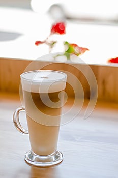 Beautiful tall transparent glass containing latte coffee drink on table in cafe in front of window with natural light