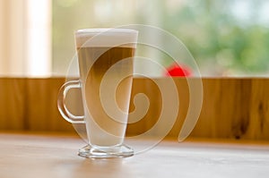 Beautiful tall transparent glass containing latte coffee drink on table in cafe in front of window with natural light