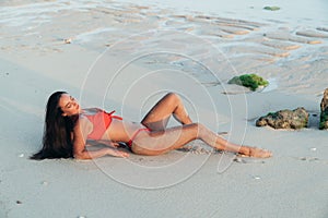 Beautiful tanned brunette girl in red swimsuit lying on sandy beach. Sexy model posing near ocean