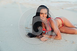 Beautiful tanned brunette girl in red swimsuit lying on sandy beach. Sexy model posing near ocean