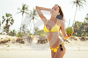 Beautiful tanned girl in trendy yellow bathing suit sunglasses stands with coconut in her hands on beach tropical island