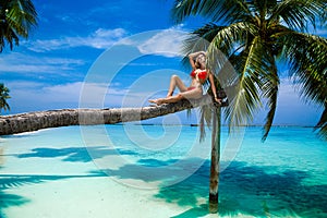 Beautiful tanned woman in colorful bikini is sitting on palm on tropical beach on Maldives island. Sexy female model relaxes on