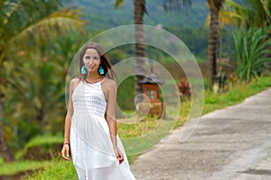 Beautiful tanned woman in white dress posing standing on the road. In the background are palm trees and other tropical vegetation