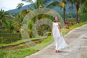 Beautiful tanned woman in white dress posing standing on the road. In the background are palm trees and other tropical vegetation