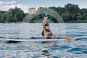 beautiful tattooed woman with blue hair sitting on sup board