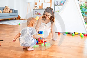 Beautiful teacher and toddler playing with building blocks around lots of toys at kindergarten