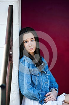 A beautiful teen brunette girl with a serious look leaning against a wall in thought with a pink red background