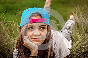 Beautiful teen girl with big eyes in a baseball cap lies on the grass and looks away