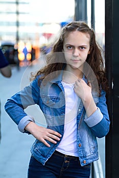 Beautiful teen girl in denim clothes stands leaning on a glass building on the city street