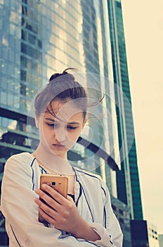 A beautiful teen girl stands on a background of modern buildings and holds a smartphone in her hands.