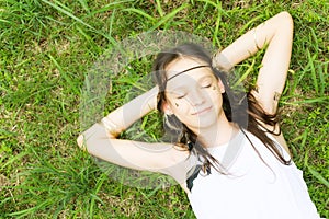 Beautiful teen girl in white dress lying on green grass, smiling, eyes closed. Top view. Boho style portrait