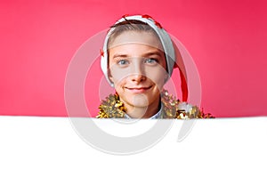 beautiful teen in Santa hat and with tinsel on neck, posing behind white panel isolated on red background. Teen baby holding an e