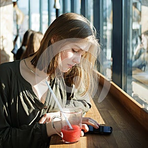 Beautiful teen student girl ?oncentrated attention using smartphone for internet surfing at street cafe sitting near big window ho