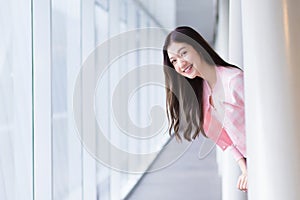 Beautiful teenage Asian girl wears a white with pink shirt and smiles cheerfully while she stands near the glassed window on a