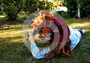a beautiful teenage girl in a big red hat is lying on the grass with a multicolored bouquet of flowers.