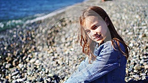 A beautiful teenage girl in a denim jacket sits on a pebble seashore
