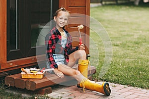 Beautiful teenage girl in denim overalls and a plaid shirt holds a roller in her hands and smiles. A happy family. Home