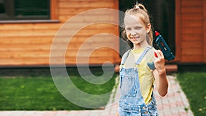 A beautiful teenage girl in denim overalls and a yellow T-shirt holds a drill in her hands and smiles. A happy family. Home