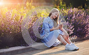 Beautiful teenage girl listening to music in headphones from smartphone. young happy model sitting outdoors in park flowers on