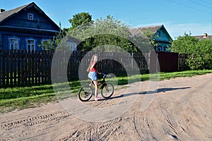 beautiful teenage girl with long blonde hair is learning to ride bicycle through streets of summer village on sunny day.