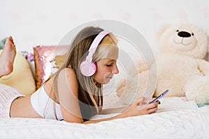 Beautiful teenage girl is relaxing in bed using a touchscreen smartphone, listening to music with a wireless headset, surfing the