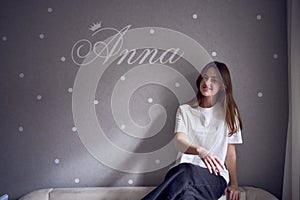 a beautiful teenage girl sits on a bed near the wall with her name written on it