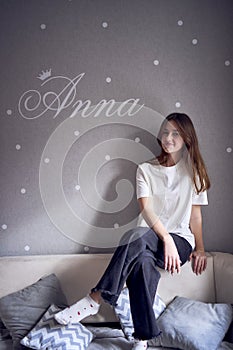 a beautiful teenage girl sits on a bed near the wall with her name written on it