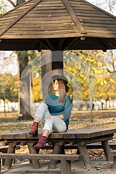 Beautiful teenage girl sitting on the park bench with black hat blue sweater and ripped jeans with blurred autumn leaves in the ba