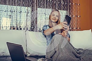 Beautiful teenage girl using laptop and taking selfie with her mobile phone, at home
