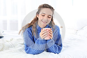 Beautiful teenage girl in warm cozy sweater with cup of hot drink on bed