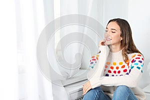 Beautiful teenage girl in warm cozy sweater looking out window at home