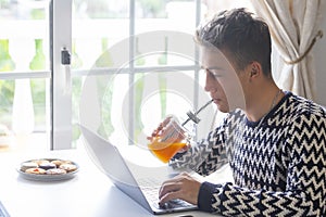 Beautiful teenager drinking an orange juice using laptop at home in front of the window -  Millenial generation enjoying