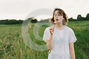 Beautiful teenager girl on the field in green grass and blowing dandelion.