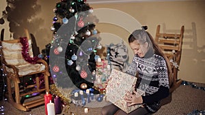 Beautiful teenager girl near a Christmas tree opens a gift box with a New Year`s gift from Santa Claus
