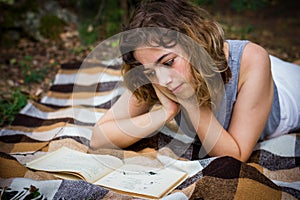 Beautiful teenager girl reading a book lying on blanket on green grass at the forest