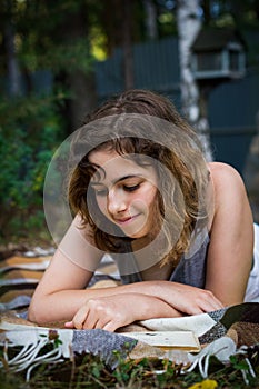 Beautiful teenager girl reading a book lying on blanket on green grass at the forest