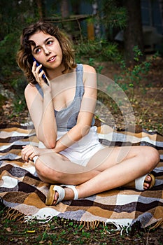 Beautiful teenager girl talking by phone and at the forest. Young girl sitting on blanket at grass