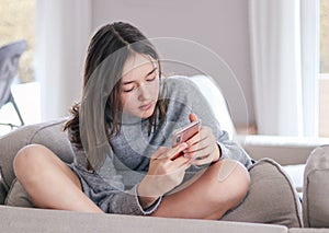 Beautiful teenager girl in warm pullover sitting on sofa pilow in funny pose with crossed legs looking at phone in her hands chatt