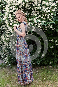 Beautiful tender blonde girl in a long dress with evening hairstyle standing in the garden near a flowering aromatic tree