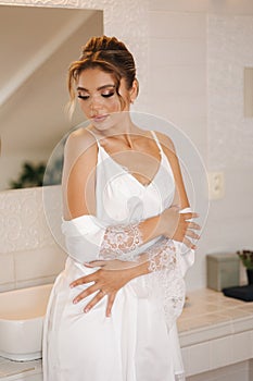 Beautiful and tender bride in white silk robe standing in luxury bathroom. Female preparing for wedding