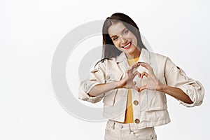 Beautiful tender girl shows heart sign, woman make I love you gesture and smiling cute, express her feelings, standing