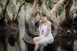 beautiful Thai asian woman in white dress local tradition costume in the name is Nakee, sitting on tree stump in the lake in the